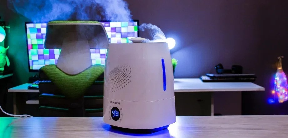 Увлажнитель Polaris PUH 5906Di белого цвета стоит на деревянной поверхности перед рабочим стулом и столом с компьютером